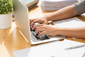 Person typing on a laptop at a wooden desk with papers and a potted plant, indicating a work or study environment.