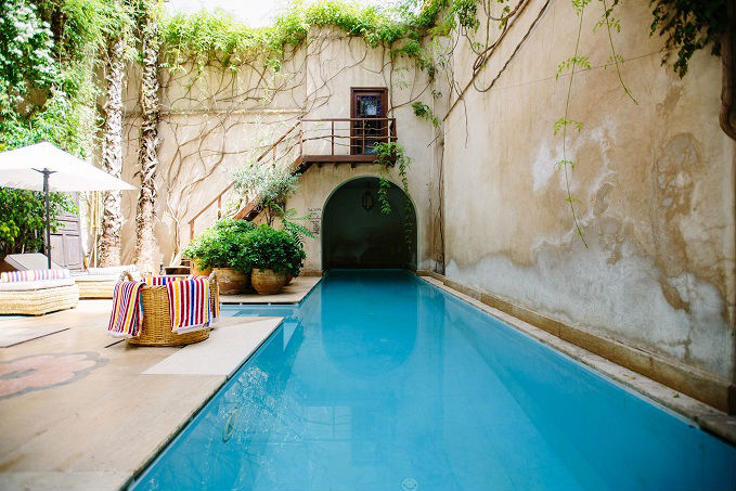 A serene courtyard with a narrow swimming pool, surrounded by weathered walls with climbing vines, potted plants, and lounge chairs with colorful cushions under a white umbrella.