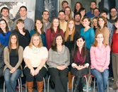 A group of people arranged in four rows, with the back rows standing and the front rows seated, dressed in casual to business casual attire, smiling for a group photo in an indoor setting.