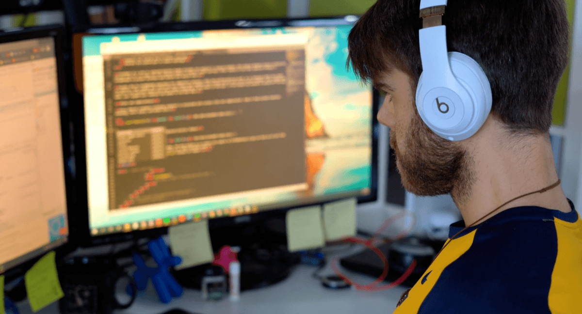 A person wearing headphones is working on a computer with multiple monitors displaying code, surrounded by a cluttered desk with sticky notes and tech accessories.