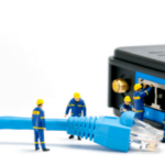 Miniature figures dressed as technicians working on a large Ethernet cable and network switch, with one figure holding a small golden globe, against a white background.