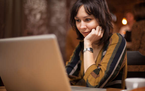 A woman with short hair, wearing a striped shirt and a watch, is looking at her laptop screen with interest while sitting in a warmly lit indoor space.