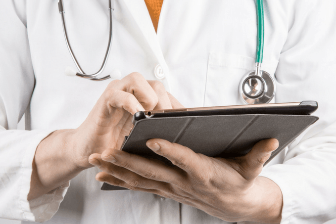 Two healthcare professionals in white coats, one with a stethoscope, are looking at and interacting with a tablet.