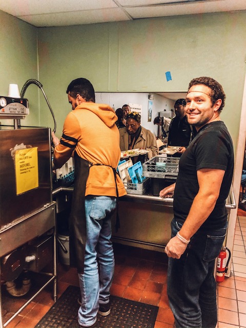 A man smiling at the camera in a commercial kitchen with other individuals working in the background.