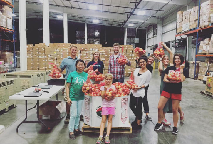 A group of nine diverse individuals posing with bags of red apples in a warehouse, likely participating in a food distribution activity.