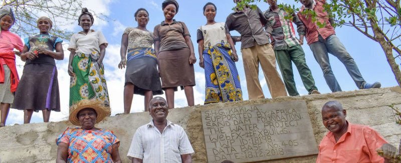 A group of men and women posing on a newly built sand dam project in a rural setting, with a sign indicating support from The Water Project, database number, and completion date.