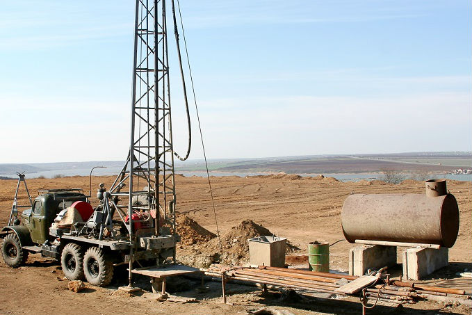 A drilling rig mounted on a truck chassis at a rural construction site with a cylindrical tank and various equipment scattered around.