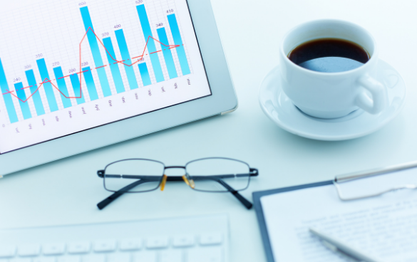 A tablet with a data chart on the screen, a cup of coffee, eyeglasses, and a clipboard with paper and a pen on a desk.