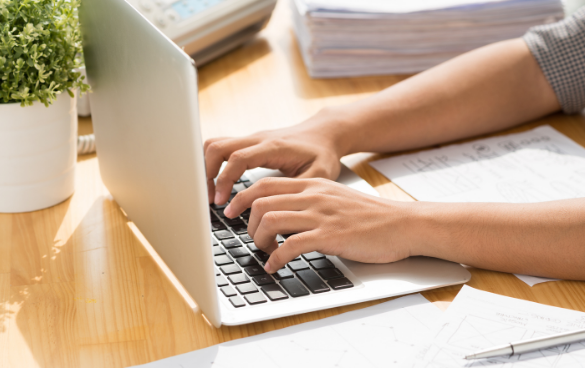 Close-up of a person's hands typing on a laptop keyboard with papers and a calculator nearby on a wooden desk.