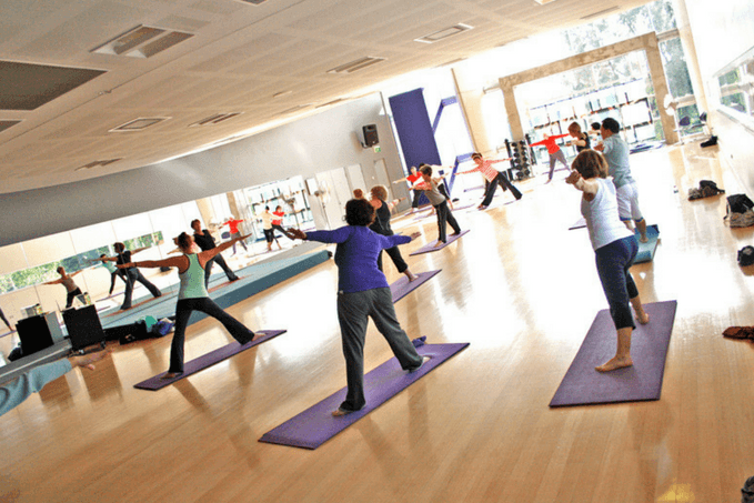 A group of people participating in a fitness class, standing on purple yoga mats and following an instructor's lead in a bright, mirrored room.