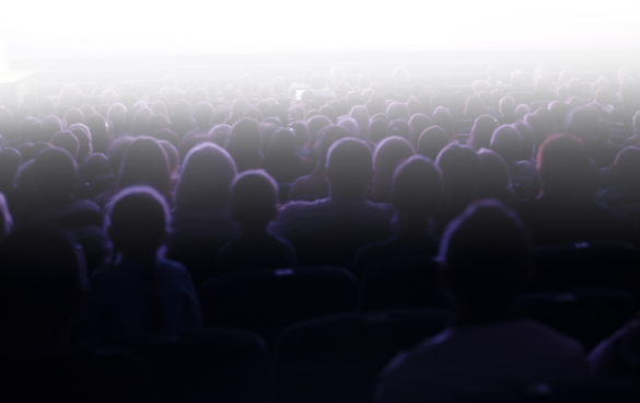 Blurred silhouette of an audience in a dark theater facing a bright screen or stage.