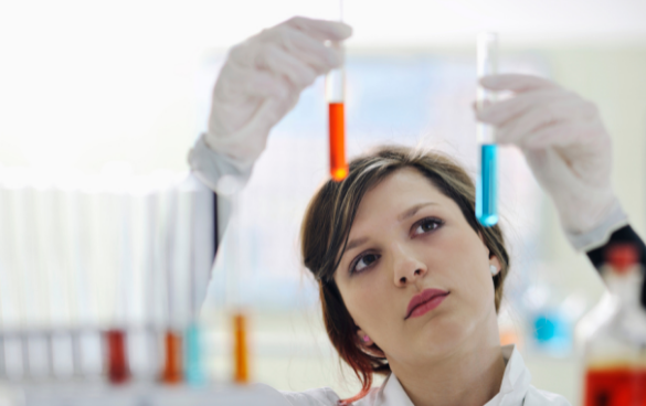 A female scientist in a lab coat and gloves thoughtfully examining the contents of two test tubes, one with orange liquid and the other with blue, in a laboratory environment.