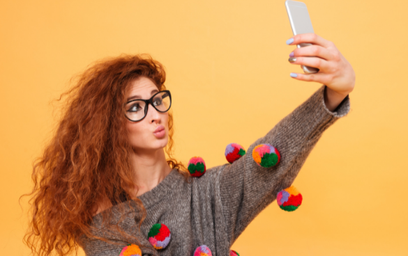 A woman in a brown sweater poses while taking a selfie with her phone