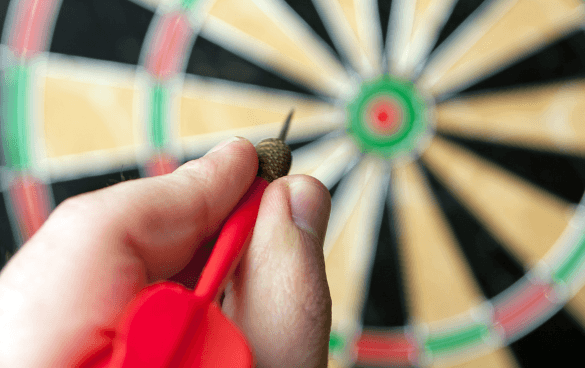 Close-up of a hand holding a red dart aimed at a blurred dartboard in the background.