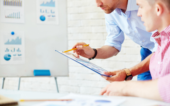 Two professionals discussing a document with charts and graphs in an office setting, with one person pointing at the document and explaining, while the other listens attentively.