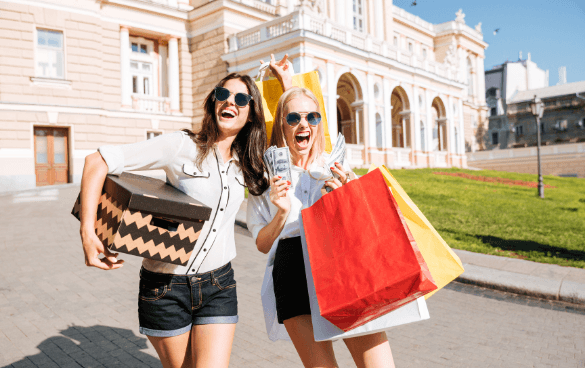 Two joyful women with sunglasses holding shopping bags and cash, celebrating a shopping spree in front of a classical building on a sunny day.