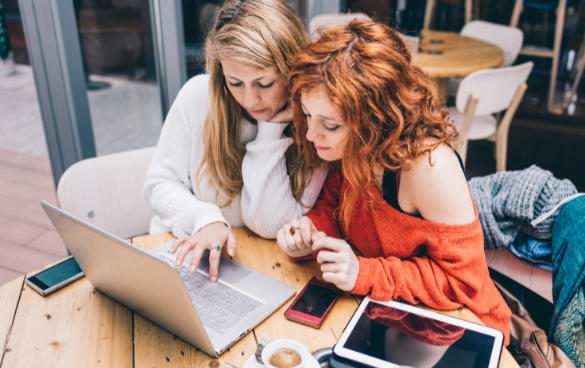 Two women sitting at a wooden table in a café, looking at a laptop screen with a smartphone, tablet, and a cup of coffee on the table.