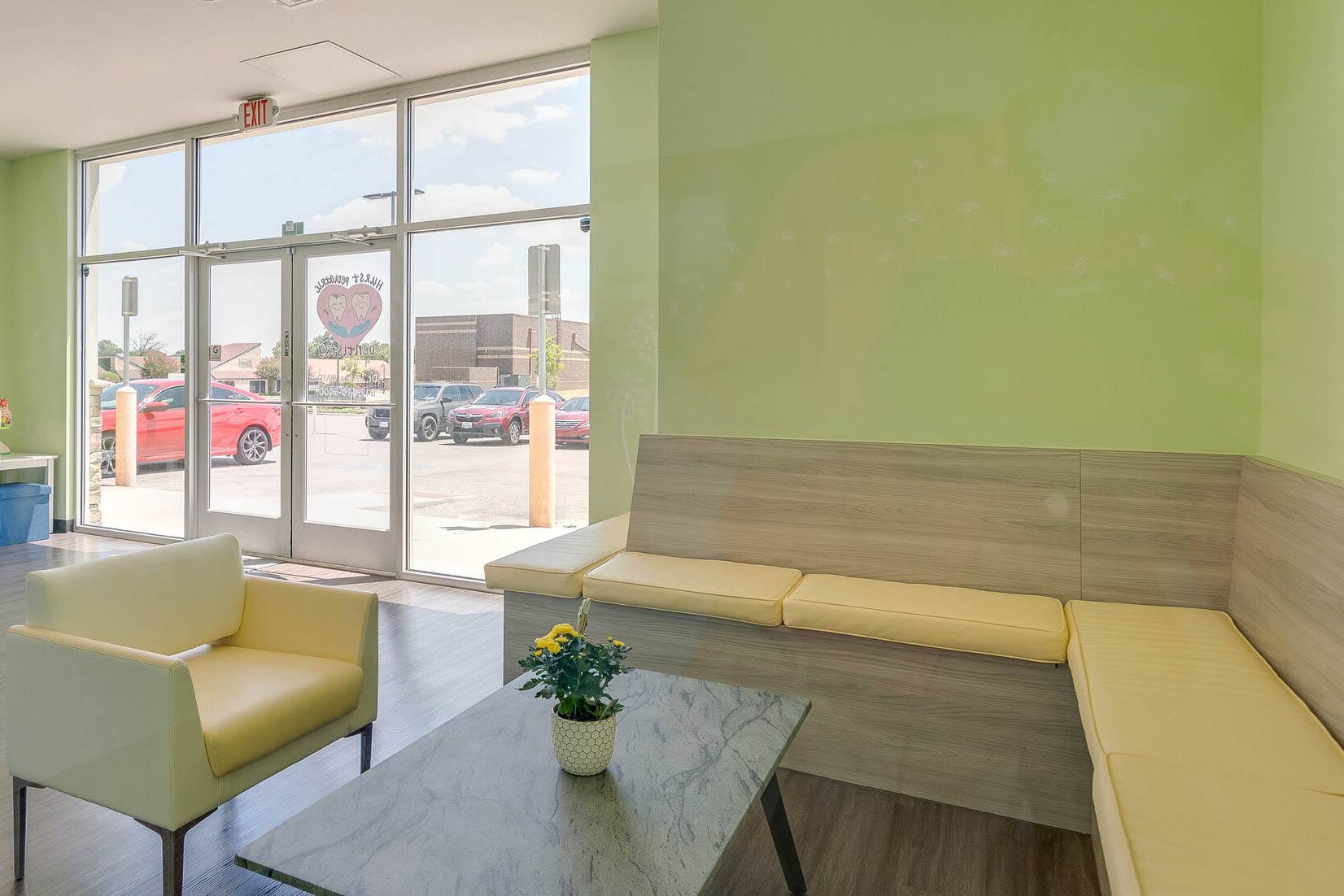 Bright interior of a room with light green walls and a pattern of white flowers, featuring a yellow bench-style seat, a yellow armchair, and a marble-patterned table with a potted yellow flower. Glass doors lead to a parking lot visible outside.