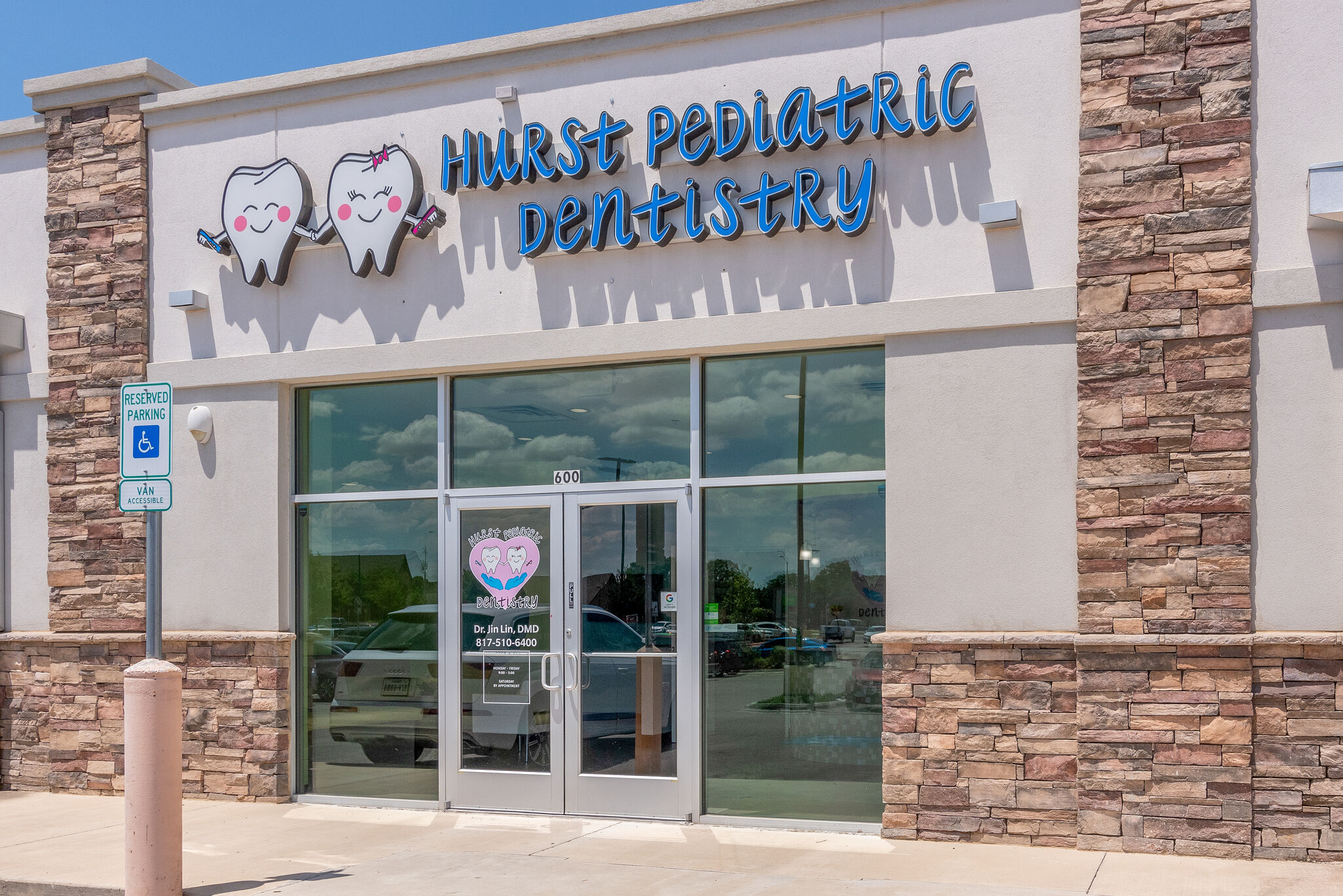 Storefront of Hurst Pediatric Dentistry with a sign featuring cartoon teeth, glass doors with business logo, and a reserved parking sign for van accessible spaces.