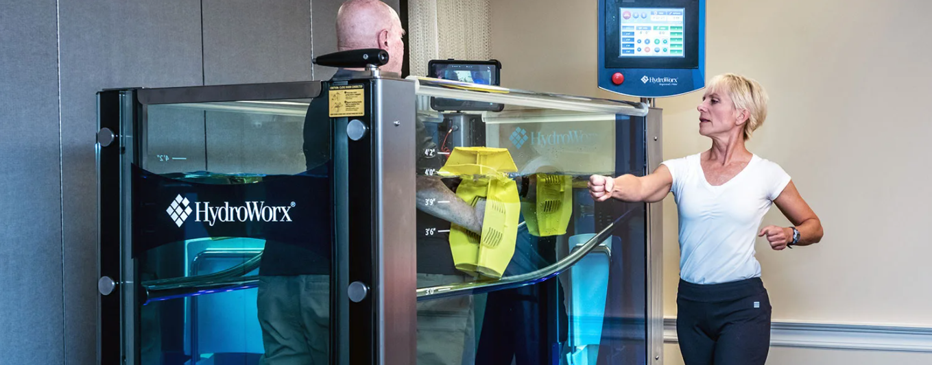 A man using an aquatic treadmill in a HydroWorx chamber while a woman runs alongside, coaching him in a rehabilitation facility.