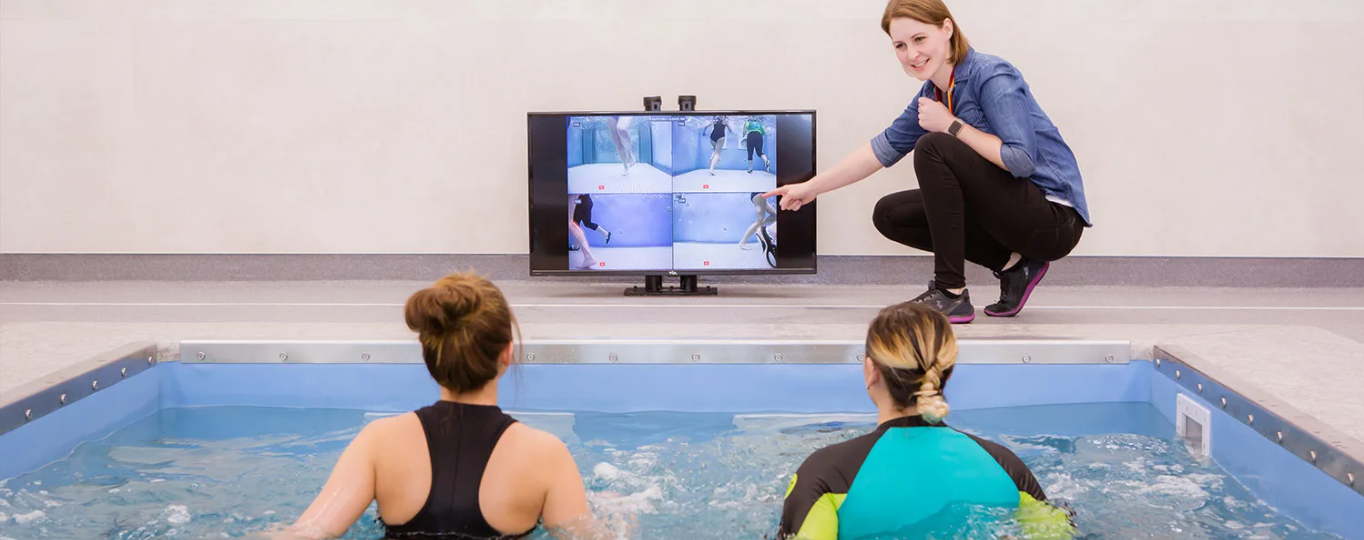 Two people in a swim training pool watching a multi-view screen of a swimmer, with an instructor outside the pool pointing at the screen and giving a thumbs-up.