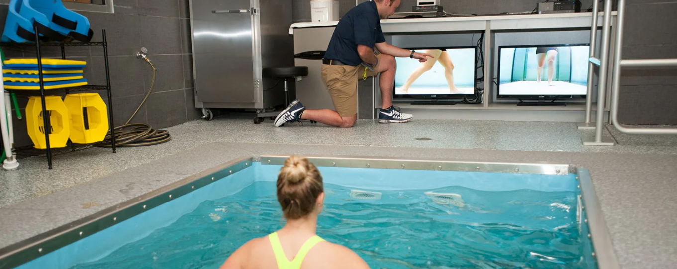 A woman in a hydrotherapy pool watches as a man kneeling beside the pool points to a screen displaying her swimming technique, with multiple monitors showing different angles of her swim stroke in a professional training facility.