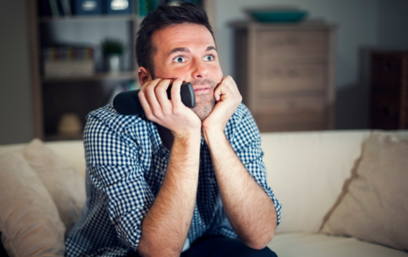 Man sitting on a couch holding a remote control, looking excited or anticipatory while watching television.