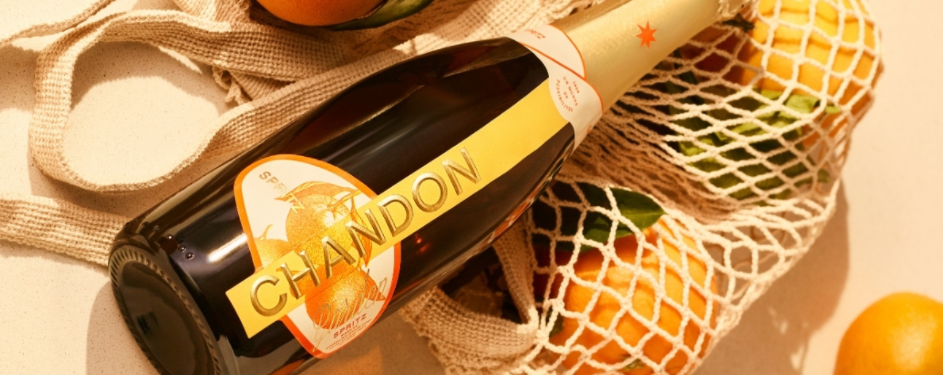 A bottle of Chandon sparkling wine with a citrus-themed label, lying next to a mesh bag filled with oranges and lemons on a beige surface.