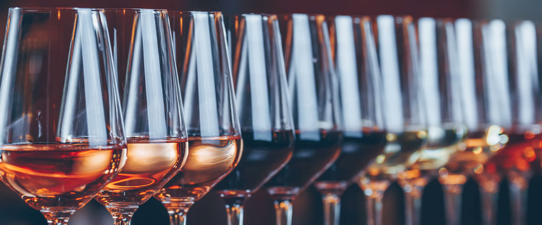 A row of wine glasses partially filled with rose wine on a reflective surface, with the focus on the first few glasses and the rest fading into a blur.