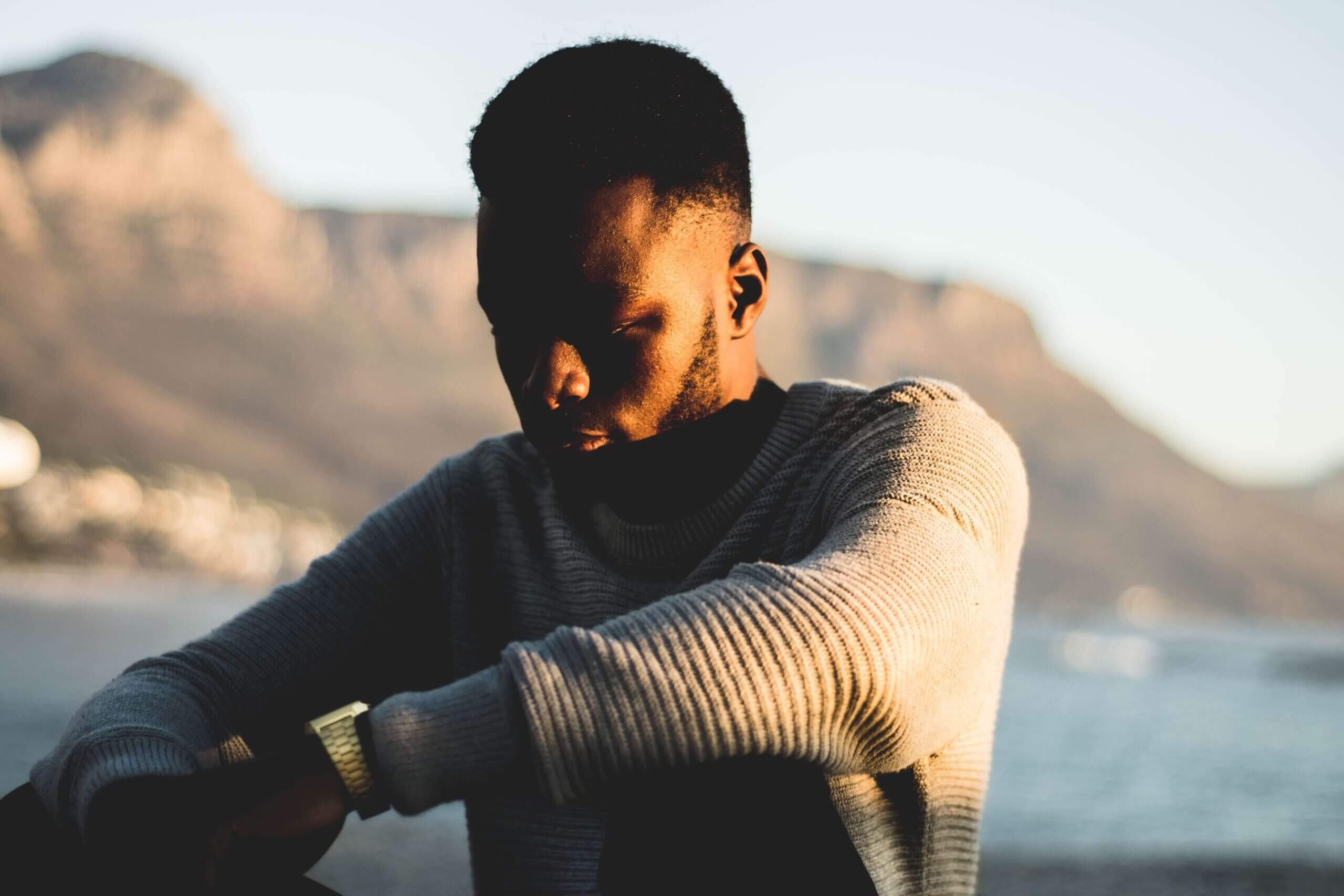 A contemplative man in a sweater with sunlight casting shadows on his face, with a blurred mountainous landscape and water in the background.