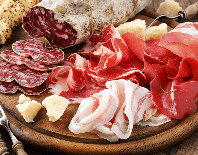 Assorted cured meats such as salami and prosciutto, along with crumbly cheese, on a wooden cutting board with a breadstick and a knife to the side.