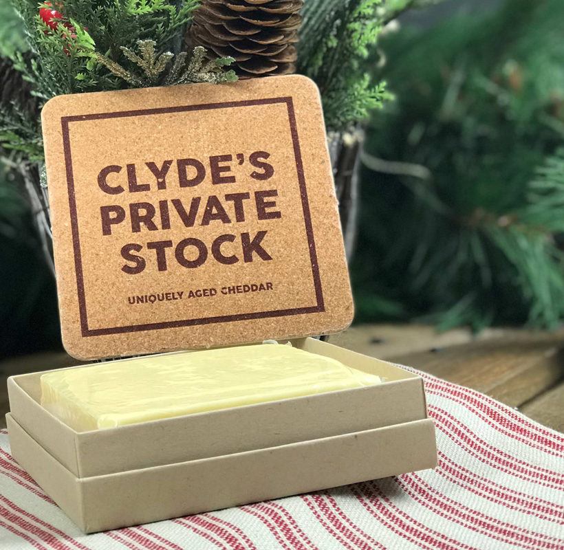 A block of cheddar cheese in an open cardboard box with a cork board label reading 'CLYDE'S PRIVATE STOCK UNIQUELY AGED CHEDDAR', set on a red and white striped cloth with pine branches in the background.