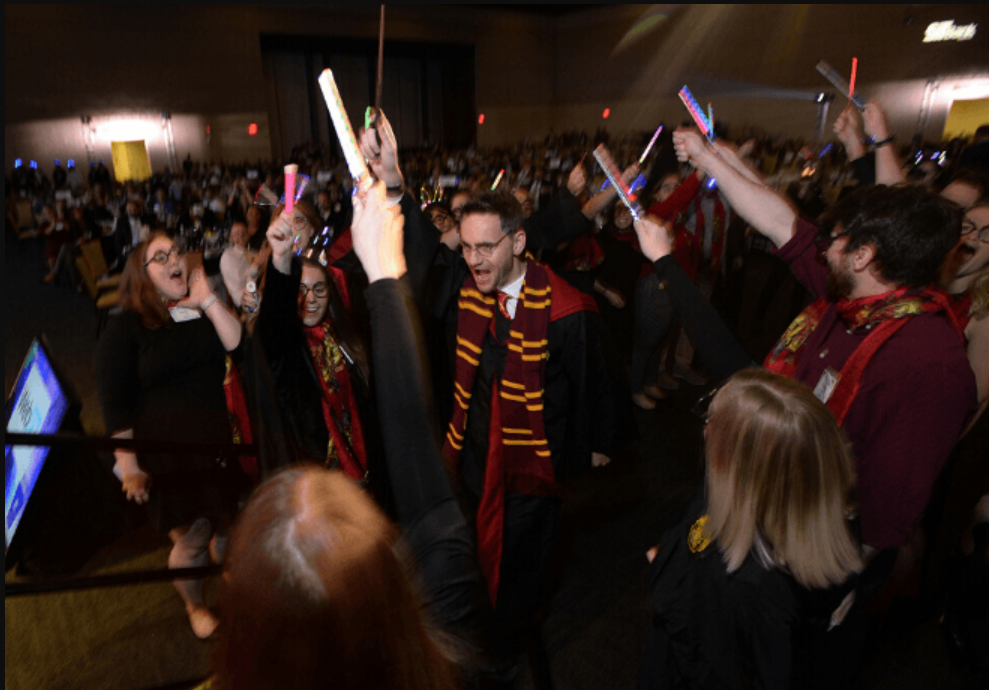 Excited fans at a Harry Potter themed event, wearing Gryffindor scarves and holding up wands and light sticks.