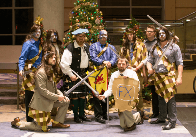 A group of people in medieval and fantasy-inspired costumes posing in front of a Christmas tree, with one person holding a cardboard sign with 'FX' written on it.