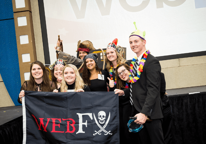 A group of people celebrating with a 'WEB F' pirate flag, wearing festive accessories, in front of a 'WINNER' backdrop at an event.