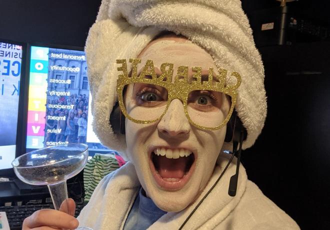 Person with a facial mask and towel on their head wearing 'CHEERS' glasses, holding a cocktail glass, and wearing a headset with a microphone, in front of a computer monitor.