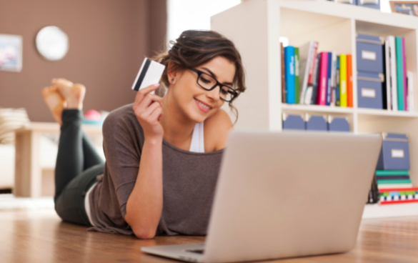 A smiling woman wearing glasses lies on her stomach on the floor, looking at a laptop and holding a credit card, with a bookshelf in the background.