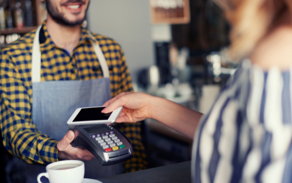 A smiling server in a checkered apron accepts a contactless payment via smartphone from a customer at a coffee shop counter, with a cup of coffee visible in the foreground.