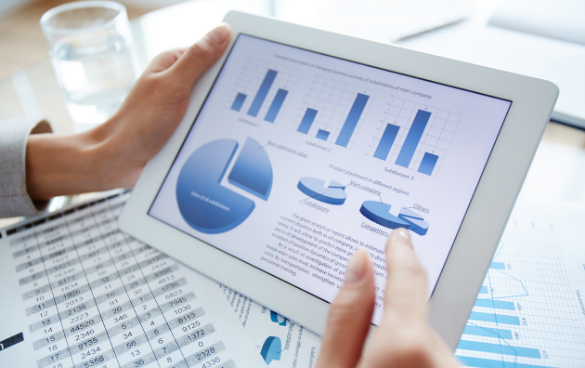 Person holding a tablet with data charts and graphs on the screen, with a printed spreadsheet visible on the table.