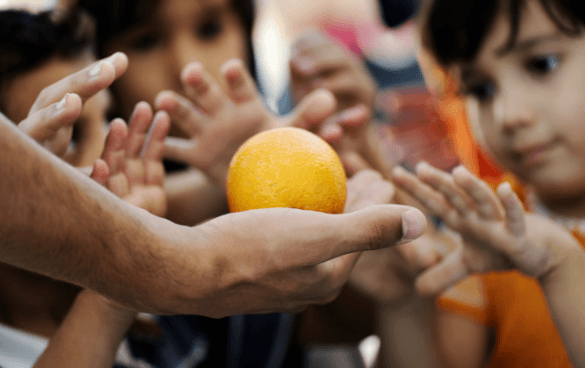 An adult hand presenting an orange to several children with outstretched hands, eager to receive the fruit.