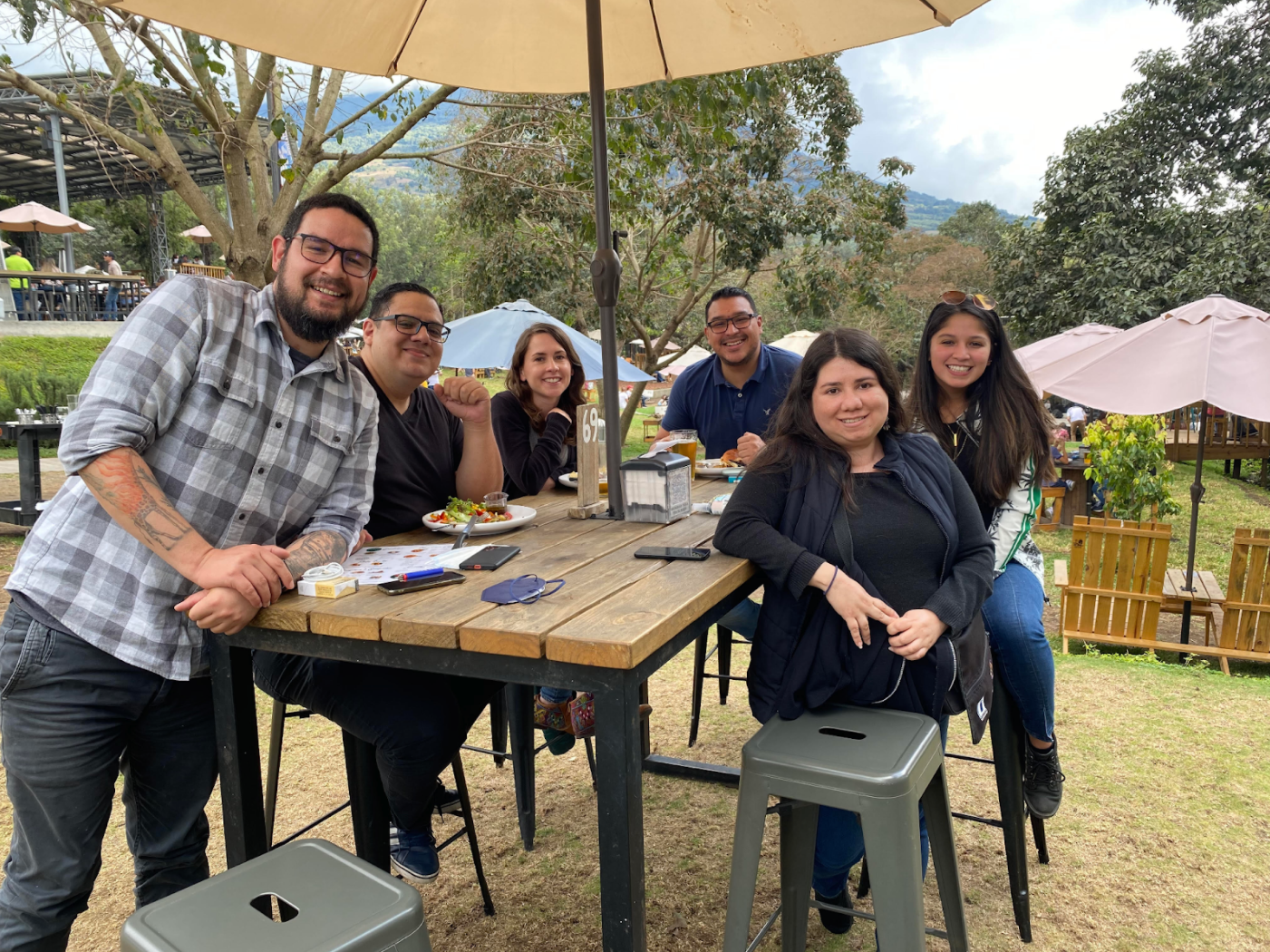 A group of six people smiling and posing for a photo around a wooden table at an outdoor dining area with food and drinks on the table, and other diners and greenery in the background.