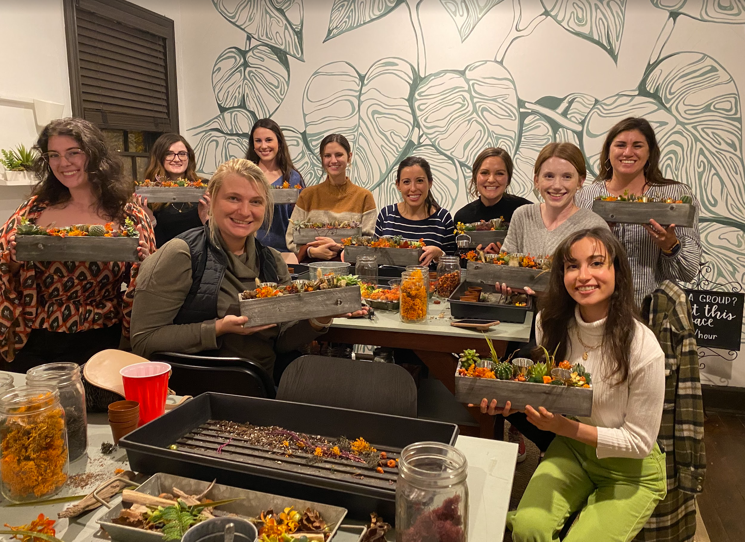 A group of eight women holding handmade succulent planters, standing in front of a leafy mural in a crafting workshop, with a joyful and creative ambiance.