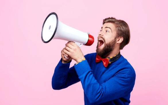 Enthusiastic bearded man in a blue sweater and red bow tie shouting into a megaphone against a pink background.