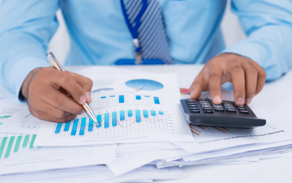 A person in a light blue shirt and striped tie analyzing financial charts and using a calculator at a desk.