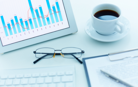 A business workspace with a tablet showing a graph, eyeglasses, a cup of coffee, a keyboard, and a clipboard with paper and pen.