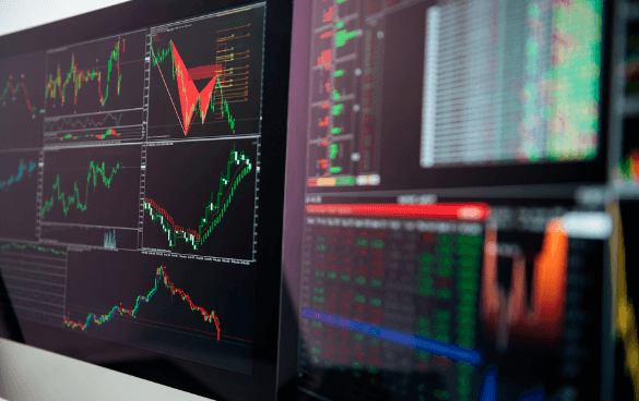 Multiple computer monitors displaying financial charts and trading data with colorful graphs and numerical information.