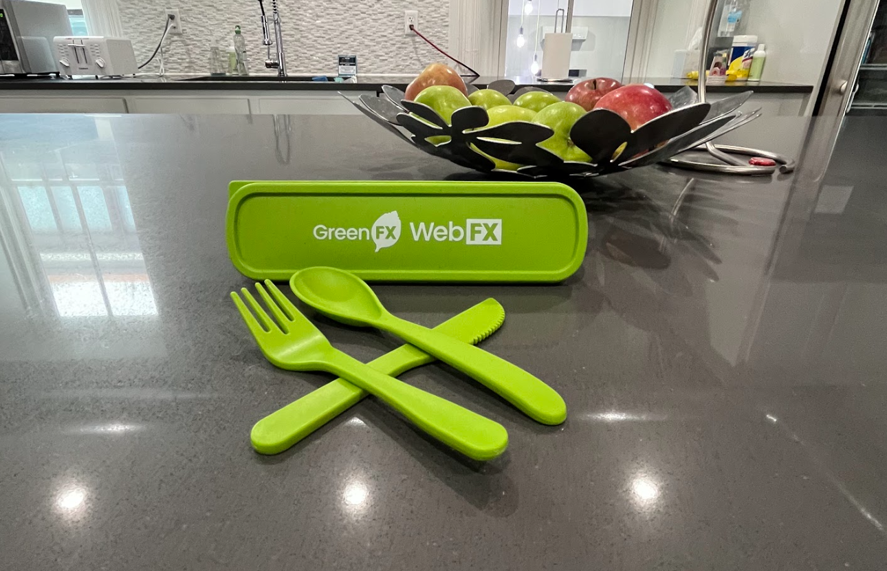 Bright green reusable cutlery set with a fork, knife, and spoon, alongside a green case with 'GreenFX WebFX' text, on a dark grey kitchen countertop with a bowl of apples and a sink in the background.