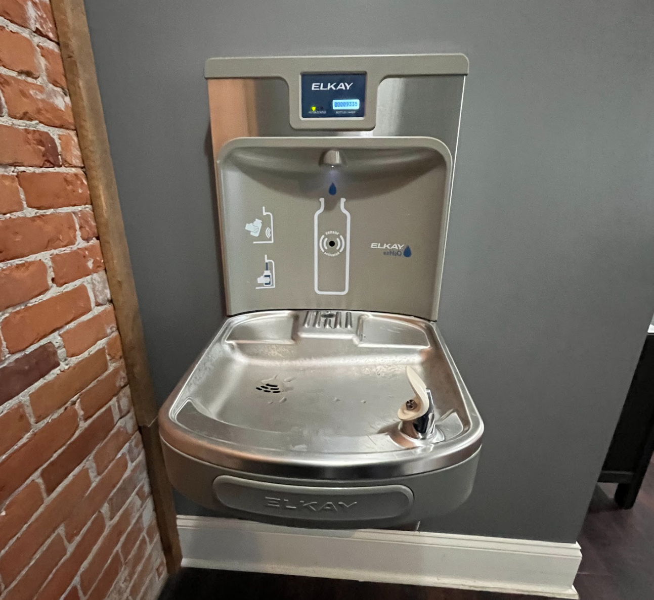 A stainless steel ELKAY water fountain and bottle filling station with a digital counter, mounted on a wall with exposed brick on one side.