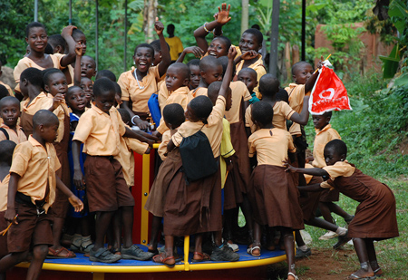 A group of children in brown and yellow school uniforms playing on a merry-go-round, waving and smiling in a green outdoor setting.