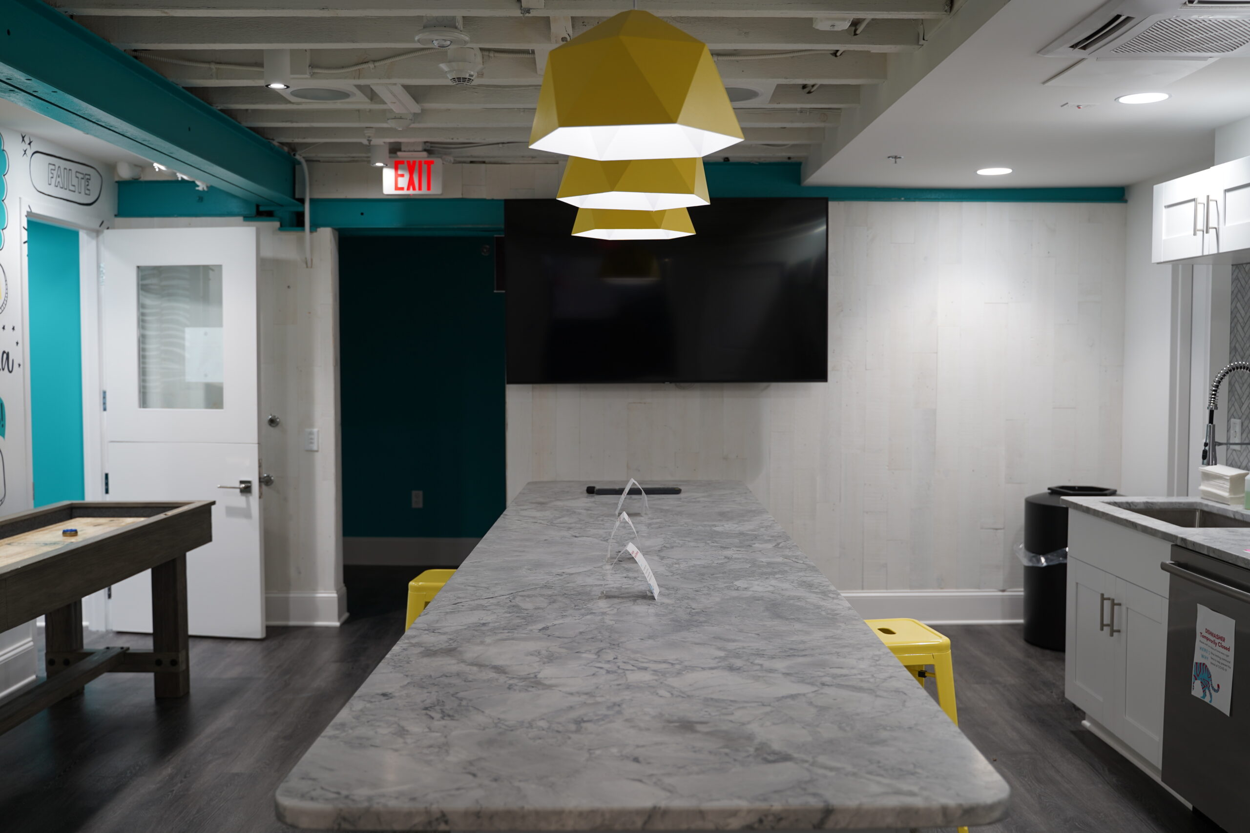 Modern break room with a long marble countertop, geometric pendant light, wooden game table, white cabinetry, and a mounted flat-screen TV. The room features teal and white walls with dark hardwood flooring.